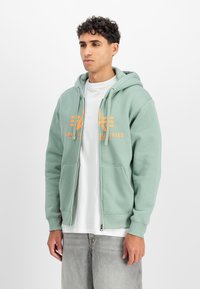 Férfi világoszöld, cipzáros kapucnis felsőt visel narancssárga logóval, fehér pólót és világosszürke farmert, fehér háttér előtt áll.