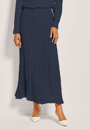 Vrouw draagt een marineblauwe lange geplooide rok, bijpassende top met lange mouwen en witte puntige hakken, staand tegen een lichte achtergrond.