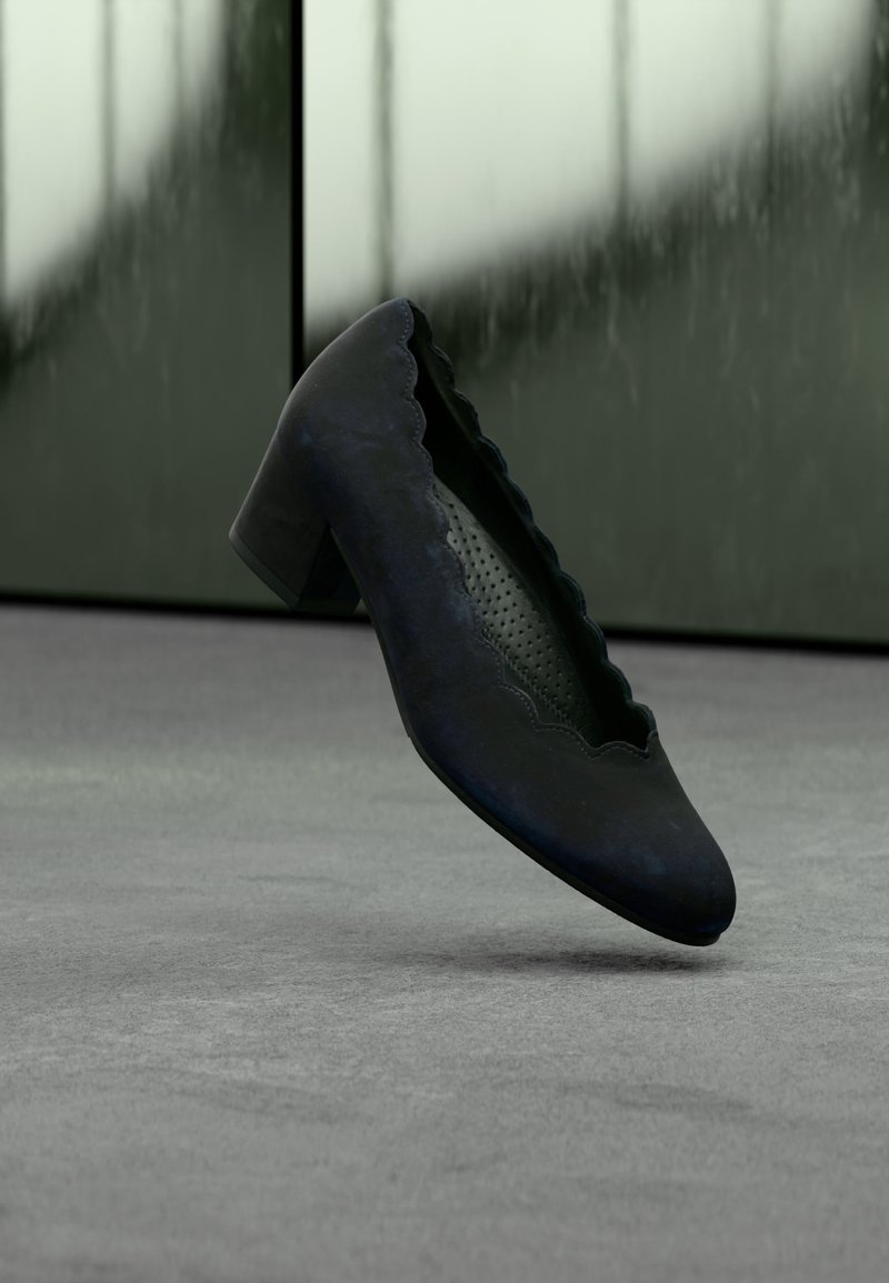 Chaussure noire à petit talon avec bords festonnés et semelle intérieure perforée, flottant au-dessus d’une surface grise avec un fond vert flou.