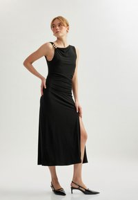 Schwarzes ärmelloses Kleid aus softem Stoff mit drapiertem Ausschnitt, seitlichem Schlitz und goldenen Hardware-Akzenten an der Schulter.