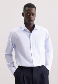 Lichtblauwe button-up shirt met een subtiel ruitpatroon, enkele borstzak en lange mouwen. Gecombineerd met donkere, geplooide broek.