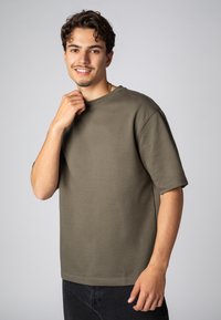Olive green cotton t-shirt with a crew neck and short, loose sleeves. Features a straight cut and soft texture. Worn with dark pants.