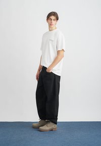 Biała bawełniana koszulka z małym graficznym nadrukiem na piersi, w połączeniu z luźnymi czarnymi spodniami i beżowymi butami, na jednolitym tle.