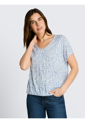 Femme aux longs cheveux bruns portant un chemisier blanc à manches courtes avec un imprimé floral bleu et un jean bleu foncé, posant avec une main sur le cou.