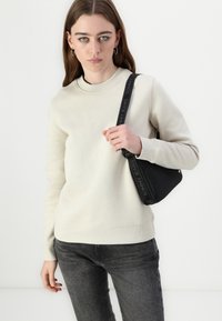 Krémová mikina s rebrovaným okrajom okolo krku, v kombinácii s tmavošedými džínami. Doplnková čierna taška na rameno Tommy Jeans s logom na popruhu.