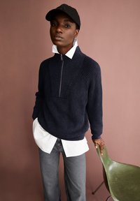 Marineblauer Rippstrickpullover mit hohem Kragen und halbem Reißverschluss, über einem weißen Kragenhemd getragen, kombiniert mit grauen Jeans und einer schwarzen Mütze. Grünem Stuhl.