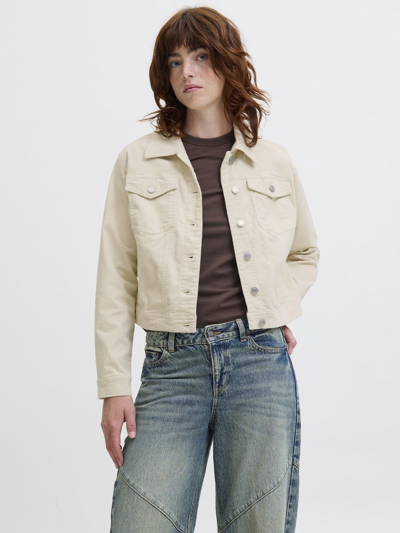 Olkapäihin ulottuvat aaltoilevat punaiset hiukset omaava nainen pukeutuneena beigeen napitettuun takkiin, joka on päällä tummaa ruskeaa toppia, sekä leveälahkeisiin haalistuneisiin sinisiin farkkuihin, seisoo yksiväristä valkoista taustaa vasten.