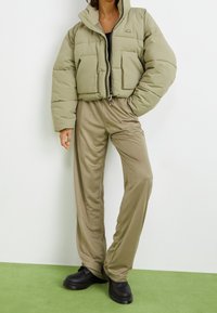 Short puffer jacket in light green color, featuring a high collar and front pockets, paired with beige, loose-fitting pants and black shoes.