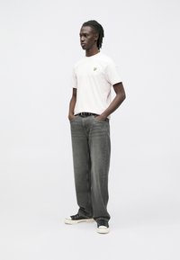 Lichtroze T-shirt met korte mouwen en een geel logo, gecombineerd met grijze jeans met rechte pijpen. Zwarte sneakers met witte zolen maken de look compleet.