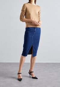 Mujer vistiendo una blusa de manga larga color camel, una falda de mezclilla oscura hasta la rodilla con abertura frontal y unos tacones negros de punta con correas en los tobillos.