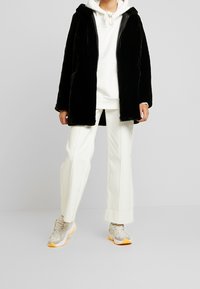 Abrigo de piel sintética negra con capucha, combinado con una sudadera blanca y pantalones de pierna ancha color crema. Las zapatillas presentan patrones grises con acentos naranjas.