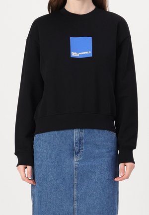 Persona con una sudadera negra con un logo cuadrado azul que dice "KARL LAGERFELD JEANS" y una falda vaquera azul.