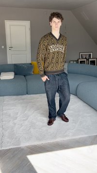 Maglione con un motivo marrone e oro, a scollo a V, abbinato a jeans scuri larghi e scarpe marroni, in piedi su un tappeto chiaro.