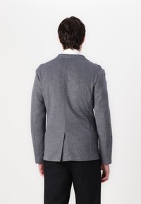 Γκρίζο σακάκι με υφή, με δομημένο γιακά, πίσω ανοίγμα και δύο πλαϊνές τσέπες, σε συνδυασμό με μαύρο παντελόνι. Πίσω όψη του ενδύματος.