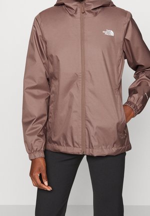Veste marron zippée avec capuche, poignets élastiques et logo The North Face sur la poitrine gauche, portée par-dessus un pantalon noir.