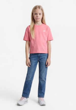 Νεαρή κοπέλα όρθια, φοράει ροζ μπλούζα, μπλε τζιν και λευκά αθλητικά με ροζ κορδόνια, με φόντο λευκό.