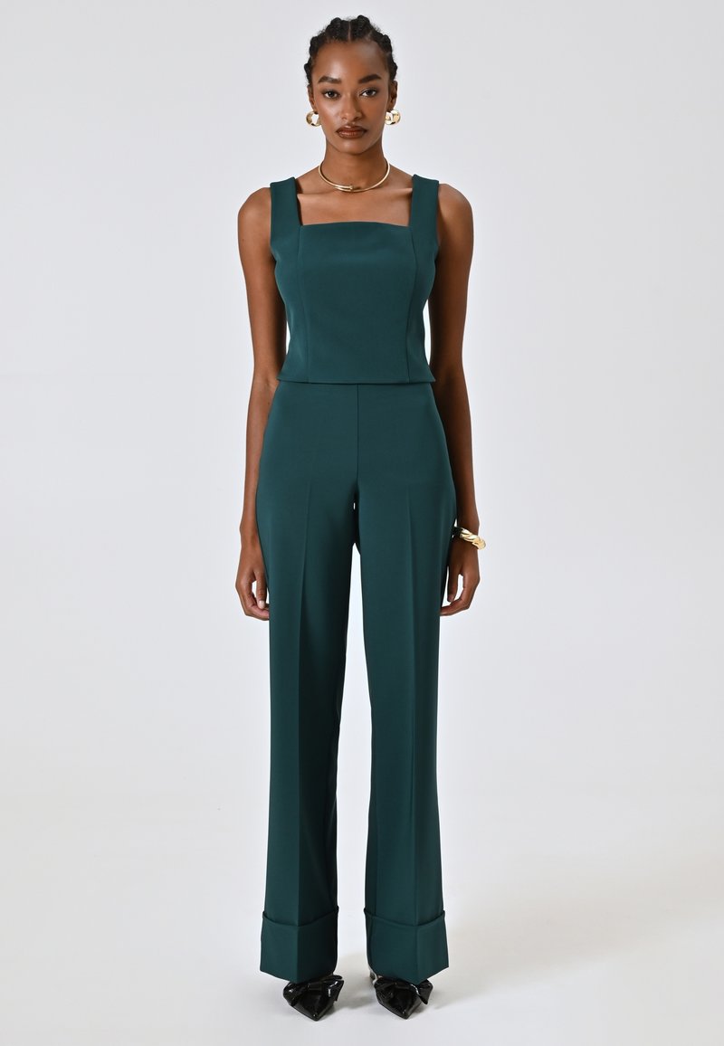 Tutina teal con scollo quadrato, spalline larghe e gambe svasate, realizzata in tessuto morbido. Si chiude con risvolti ai polsini e tacchi neri appuntiti.