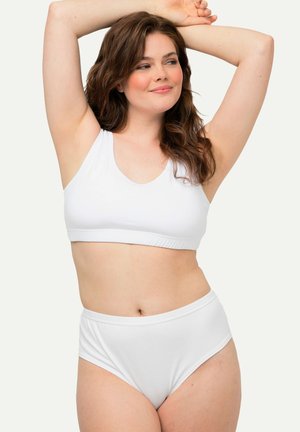 Woman with long brown hair wearing a white sleeveless crop top and high-waisted briefs, posing with arms raised against a plain background.