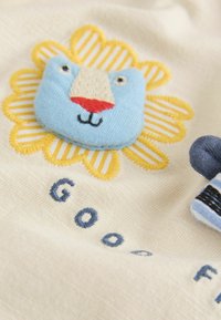 Bombažna tkanina s svetlo bež ozadjem, na kateri je vezen modri obraz leva, obdan z rumenimi cvetnimi listi in modrim napisom "GOOD."