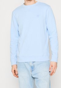 Sudadera azul claro hecha de material suave, con cuello redondo, puños acanalados y un pequeño logo cuadrado en el pecho.