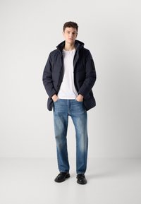 Giacca a vento blu navy con cappuccio, dettagli neri, chiusura a bottoni, indossata sopra una maglietta bianca. Abbinata a jeans azzurri chiari e scarpe nere.