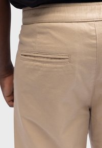 Pantalones de algodón beige con una textura suave, que cuentan con un único bolsillo trasero horizontal y detalles de costura en la cintura.