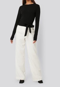 Schwarzes Langarmoberteil mit einer Knotentasche in der Taille, kombiniert mit hoch-taillierten weißen weiten Hosen und schwarzen strukturierten Schuhen.
