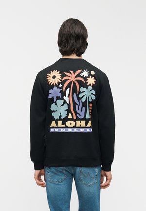 Person iført sort sweatshirt med farverigt tropisk plante- og solmotiver, tekst "ALOHA HONOLULU 1998" på ryggen.