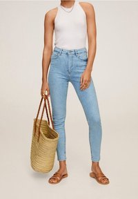 Jeans de tiro alto de color azul claro con un diseño ajustado, combinados con una blusa sin mangas blanca y sandalias planas marrones. Sosteniendo un bolso de paja trenzada.