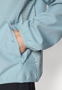 Chaqueta de color azul claro con puños elásticos y un bolsillo. Presenta un sutil patrón de puntos en el lateral. Textura de tela suave y lisa.