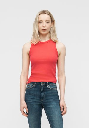 Woman with blonde hair wearing a sleeveless red top and blue studded jeans, standing against a plain light gray background.
