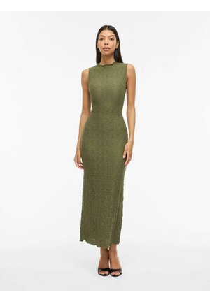 Naine seisab, kandes varrukateta, tekstuuriga oliivrohelist maksikleit kõrge kaelusega ja mustade avatud varbaga kontsadega.