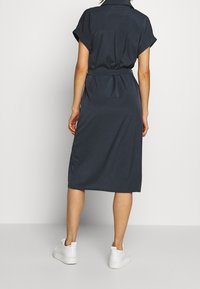 Abito camicia blu navy con maniche corte arrotolate, vita con cintura e taglio sopra il ginocchio; abbinato a sneakers bianche per un look casual.