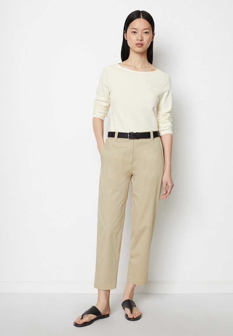 Lichtbeige langmouwtop gecombineerd met lichtkhaki broek. De riemen hebben een zwarte afwerking. Het model draagt zwarte slippers.
