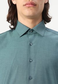 Tannengrünes Hemd mit Knöpfen aus strukturiertem Material, mit einem Kentkragen und zwei passenden Knöpfen vorne. Glattes, strukturiertes Design.