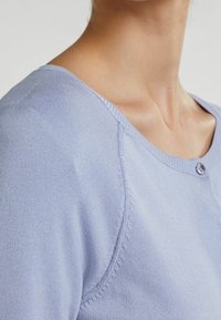 Gilet en tricot bleu clair avec un décolleté en forme de scoop, une fermeture à un seul bouton et un subtil détail côtelé le long des coutures.