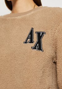 Felpa beige in pile con collo a costine e logo "AX" ricamato in nero. La texture appare morbida e soffice, ideale per il calore.