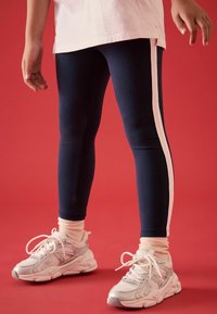 Leggings blu navali con strisce laterali bianche, abbinati a calzini alla caviglia rosa chiaro e scarpe da ginnastica grigie, su uno sfondo rosso.