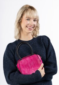 Fuchsiová kabelka s texturovaným povrchem, obdélníkovým tvarem, černými uchy a zlatými kovovými doplňky. Model ji drží blízko u tmavého svetru.