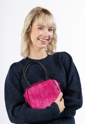 Fuchsiová kabelka s texturovaným povrchem, obdélníkovým tvarem, černými uchy a zlatými kovovými doplňky. Model ji drží blízko u tmavého svetru.