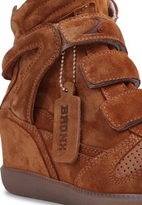 Brązowy zamszowy sneaker na koturnie z paskami na rzepy oraz skórzaną metką z napisem „BRONX” zawieszoną na metalowym łańcuszku z koralików.