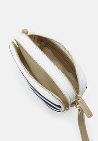 Bílá a námořnická zippovaná crossbody taška s vnitřními přihradkami; obsahuje zlaté kovové detaily a béžovou podšívku. Součástí je nastavitelné rameno.