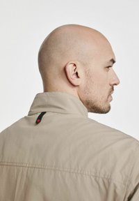 Beżowa kurtka z wysokim kołnierzem, z małą czarną i czerwoną pętelką z tyłu, gładka w dotyku, z widocznymi detalami szwów.