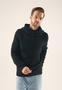 Pull noir en maille avec col cheminée et cordon de serrage, doté de poignets et d'un ourlet côtelés. Associé à un jean en denim bleu clair.