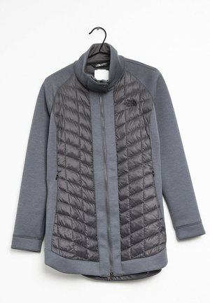 Grå quiltet North Face jakke med fuld lynlås foran, høj krave og lange ærmer hængende på en sort bøjle.
