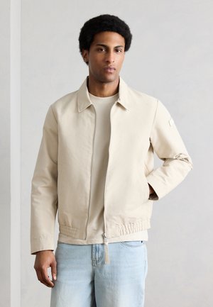 Άνδρας με κοντά σγουρά μαλλιά, φορώντας μπεζ τζάκετ με φερμουάρ και ανοιχτόχρωμο μπλε τζιν, στέκεται μπροστά σε απλό ανοιχτό φόντο.