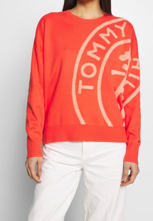 Personne portant un pull rouge vif avec un grand texte beige « TOMMY » et un motif circulaire, associé à un pantalon blanc.