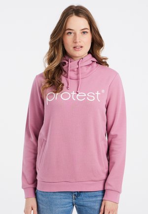 Jonge vrouw met golvend haar, gekleed in een roze hoodie met de tekst "protest" en een blauwe spijkerbroek, staand tegen een witte achtergrond.