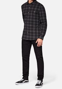 Man in zwart en grijs geruit overhemd, zwarte broek en zwart-witte sneakers, staande tegen een witte achtergrond.