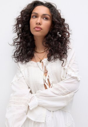 White, textured blouse with long sleeves, lace accents, and tie front. Layered gold necklaces with pendant detail.